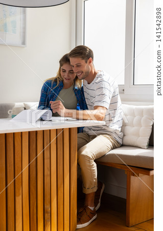 Couple leaning over papers at home dining nook reviewing documents with pen and ceramic mug Couple leaning over papers at home dining nook reviewing documents with pen and ceramic mug 141542898
