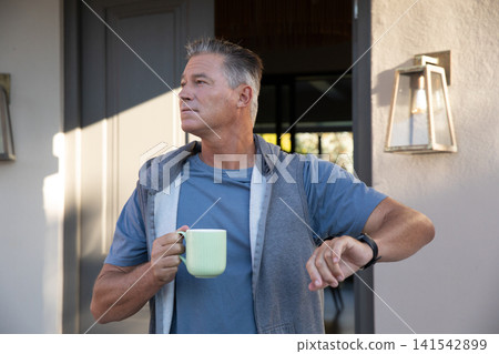 Man standing at front porch of home holding light green mug wearing smartwatch by lantern 141542899