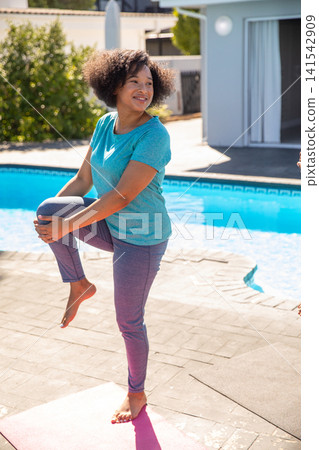 African american woman balancing one leg on pink yoga mat by pool and stretching knee toward chest African american woman balancing one leg on pink yoga mat by pool and stretching knee toward chest 141542909