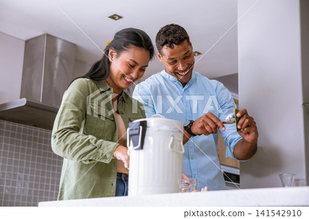 Diverse couple smiling and scooping leafy mixture from jar using small spoon at kitchen counter Diverse couple smiling and scooping leafy mixture from jar using small spoon at kitchen counter 141542910