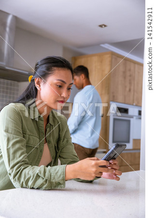 Diverse couple sitting at home kitchen countertop with woman with smartphone and man standing Diverse couple sitting at home kitchen countertop with woman with smartphone and man standing 141542911