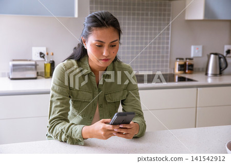 Asian woman sitting at white speckled kitchen island focusing on smartphone next to toaster Asian woman sitting at white speckled kitchen island focusing on smartphone next to toaster 141542912