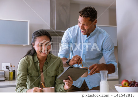 Diverse couple standing at home kitchen island looking at digital tablet, stirring mugs Diverse couple standing at home kitchen island looking at digital tablet, stirring mugs 141542913