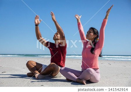 Diverse yoga partners practicing stretching in athletic wear on beach by ocean waves Diverse yoga partners practicing stretching in athletic wear on beach by ocean waves 141542914