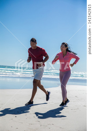 Diverse couple jogging in activewear along sandy beach under blue sky with running shoes Diverse couple jogging in activewear along sandy beach under blue sky with running shoes 141542915