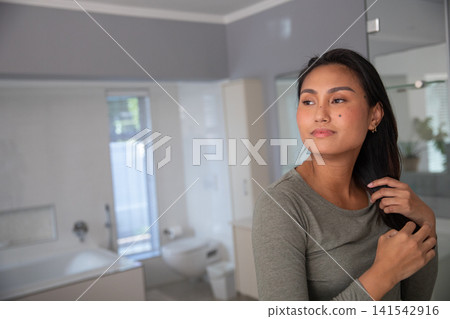 Asian woman in her 30s styling hair at vanity mirror in bathroom with bathtub, copy space Asian woman in her 30s styling hair at vanity mirror in bathroom with bathtub, copy space 141542916