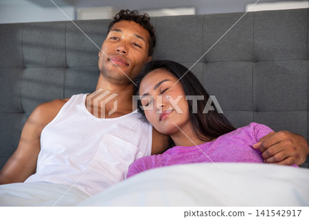Diverse couple reclining under duvet against grey upholstered headboard on bed in bedroom Diverse couple reclining under duvet against grey upholstered headboard on bed in bedroom 141542917