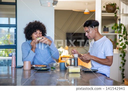 Diverse male friends biting avocado toast and spreading butter at kitchen island with French press 141542941