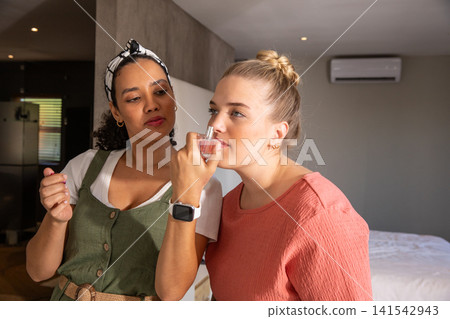 Diverse female friends standing in bedroom holding perfume bottle and checking smartwatch Diverse female friends standing in bedroom holding perfume bottle and checking smartwatch 141542943