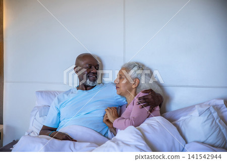 Diverse senior couple holding hands, reclining on bed with padded headboard and white pillows Diverse senior couple holding hands, reclining on bed with padded headboard and white pillows 141542944