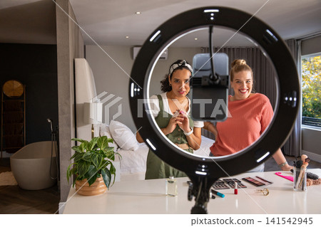 Female friends filming makeup tutorial with smartphone mount under ring light in bedroom Female friends filming makeup tutorial with smartphone mount under ring light in bedroom 141542945