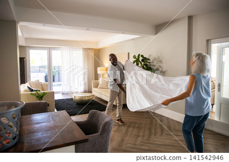 Diverse senior couple spreading white bedsheet beside plastic laundry basket in living room 141542946