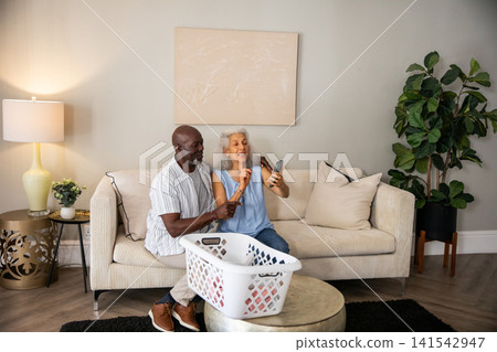 Diverse senior couple sitting on sofa at home, gesturing on smartphone near laundry basket Diverse senior couple sitting on sofa at home, gesturing on smartphone near laundry basket 141542947