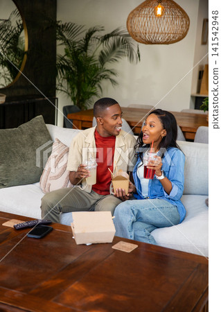 African American couple sitting on sofa in living room, holding takeout box and sipping drinks 141542948