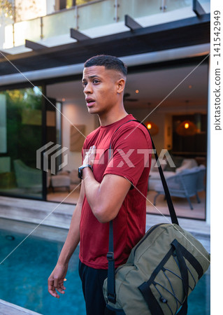 Male in mid-20s standing by pool edge on pool deck with duffel bag, smartwatch, copy space 141542949