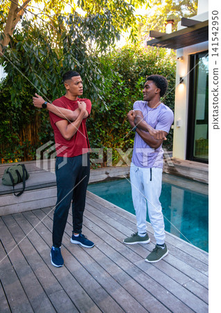 Diverse male friends stretching arms on wooden poolside deck in backyard near duffel bag and pool Diverse male friends stretching arms on wooden poolside deck in backyard near duffel bag and pool 141542950
