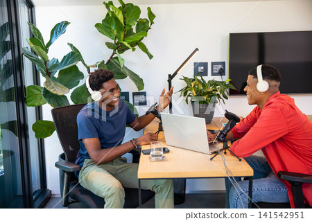 Diverse male co-hosts podcasting using microphones with laptop in modern podcast studio 141542951