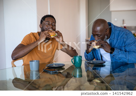 African american father and son biting sandwiches at dining table with mugs and smartphone 141542962