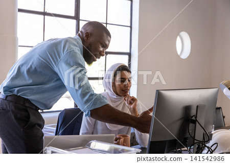 Diverse coworkers collaborating at desk in office using monitor coffee mug, safety helmet Diverse coworkers collaborating at desk in office using monitor coffee mug, safety helmet 141542986