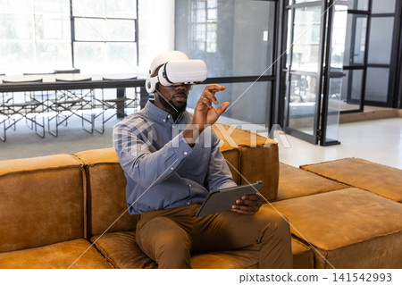 African american man wearing vr gear using tablet while sitting on leather sofa in office lounge African american man wearing vr gear using tablet while sitting on leather sofa in office lounge 141542993
