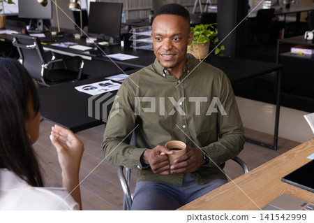 Diverse coworkers conversing across desk in open-plan office, man holding mug Diverse coworkers conversing across desk in open-plan office, man holding mug 141542999