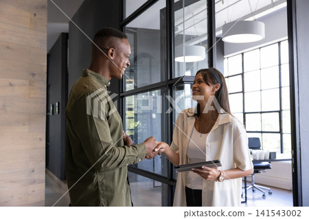 Diverse coworkers shaking hands wearing business casual attire holding tablet in office Diverse coworkers shaking hands wearing business casual attire holding tablet in office 141543002