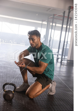 Man kneeling on gym rubber floor chalking hands by kettlebell near rig and plates with smartwatch Man kneeling on gym rubber floor chalking hands by kettlebell near rig and plates with smartwatch 141543048