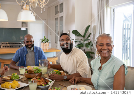 Diverse family sitting around wooden dining table in home kitchen sharing roasted chicken and salad Diverse family sitting around wooden dining table in home kitchen sharing roasted chicken and salad 141543084
