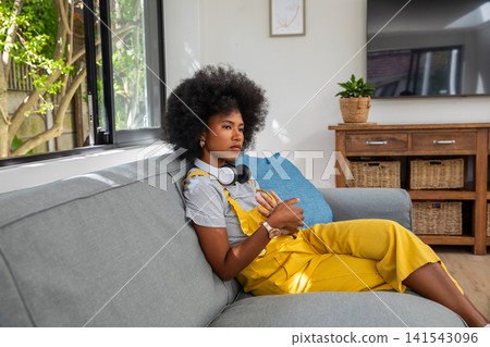 African american woman sitting on sofa at home wearing yellow overalls and headphones African american woman sitting on sofa at home wearing yellow overalls and headphones 141543096