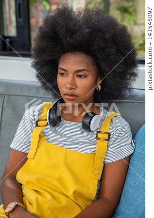 African american woman sitting on sofa wearing yellow overalls with headphones, watch, cushion African american woman sitting on sofa wearing yellow overalls with headphones, watch, cushion 141543097