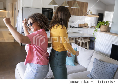 Diverse female friends dancing near gray sofa and kitchen island in open-plan living area Diverse female friends dancing near gray sofa and kitchen island in open-plan living area 141543107