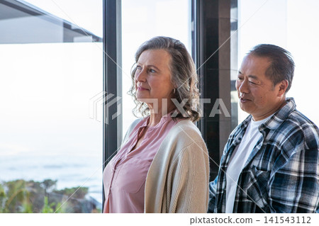 Diverse senior couple standing gazing at ocean through window inside home with coastal plants Diverse senior couple standing gazing at ocean through window inside home with coastal plants 141543112