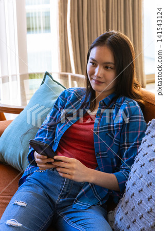 Asian woman holding smartphone on brown sofa in living room with glass side table 141543124