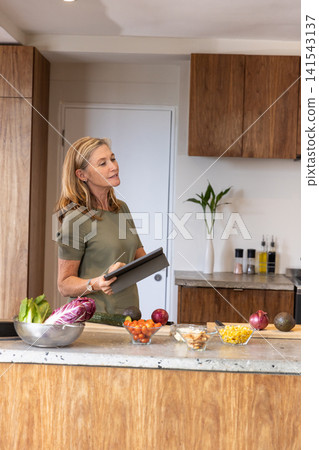 Woman holding tablet while mixing chopped vegetables in bowl in home kitchen Woman holding tablet while mixing chopped vegetables in bowl in home kitchen 141543137
