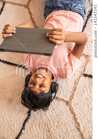 Asian preteen boy lying on beige rug on living room floor using tablet, wearing headphones 141543141
