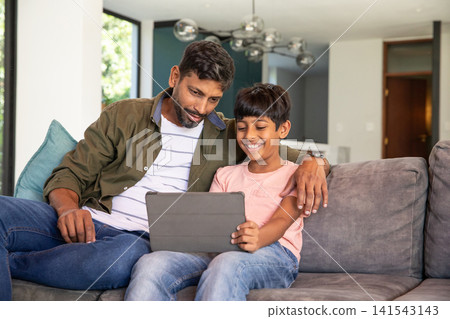 Indian father and son in casual clothes sitting on grey sofa using tablet in living room Indian father and son in casual clothes sitting on grey sofa using tablet in living room 141543143