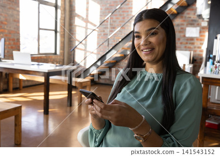 Mid adult hispanic woman holding smartphone, smiling in office by laptop, copy space Mid adult hispanic woman holding smartphone, smiling in office by laptop, copy space 141543152
