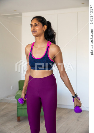 Indian woman holding purple dumbbells, wearing fitness tracker, exercising in living room Indian woman holding purple dumbbells, wearing fitness tracker, exercising in living room 141543174