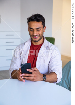 Asian man sitting at round white table with smartphone and checking wristwatch in home workspace Asian man sitting at round white table with smartphone and checking wristwatch in home workspace 141543176