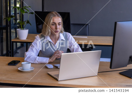 Woman in business attire working on laptop at office desk with coffee cup Woman in business attire working on laptop at office desk with coffee cup 141543194