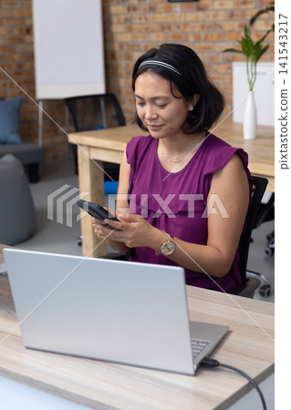 Asian woman holding smartphone, typing message at modern office desk with laptop Asian woman holding smartphone, typing message at modern office desk with laptop 141543217