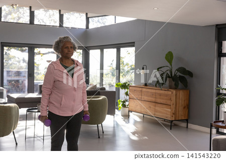 Senior african american woman lifting purple dumbbells in living room with potted plants Senior african american woman lifting purple dumbbells in living room with potted plants 141543220