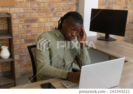 African american man sitting at desk in brick office working on laptop with smartphone African american man sitting at desk in brick office working on laptop with smartphone 141543221