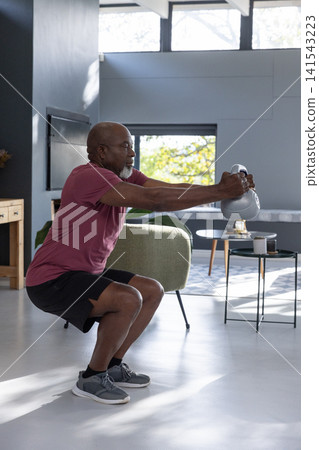 Senior african american man performing kettlebell squat in living room beside armchair Senior african american man performing kettlebell squat in living room beside armchair 141543223