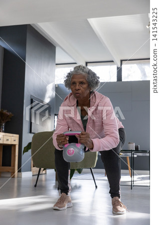 Senior african american woman performing squat holding kettlebell at home wearing sportswear Senior african american woman performing squat holding kettlebell at home wearing sportswear 141543225