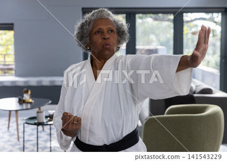 Senior african american woman practicing karate block wearing gi, black belt in living room Senior african american woman practicing karate block wearing gi, black belt in living room 141543229