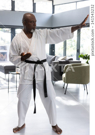 African american senior man practicing karate stance in living room with white gi, black belt African american senior man practicing karate stance in living room with white gi, black belt 141543232