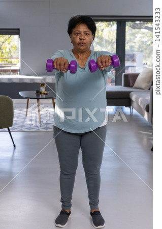 Senior indian woman lifting purple dumbbells in living room wearing top and grey leggings 141543233