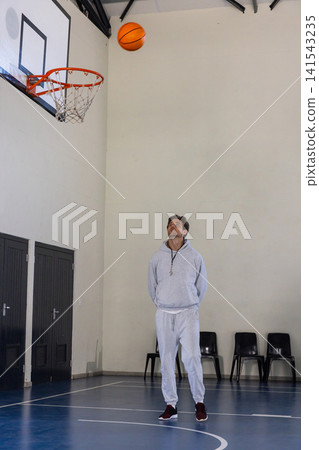 Male coach standing on basketball court wearing hoodie, whistle as basketball drops toward hoop Male coach standing on basketball court wearing hoodie, whistle as basketball drops toward hoop 141543235