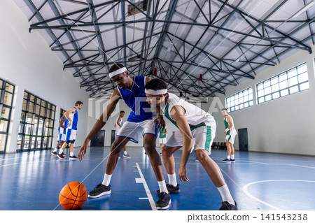 Diverse male opponents in jerseys dribbling basketball, defending on court by glass doors at gym Diverse male opponents in jerseys dribbling basketball, defending on court by glass doors at gym 141543238